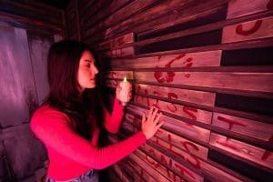 A woman in a red shirt holds a candle and touches a wooden wall marked with red handprints and letters, immersing herself in the mystery of Escapology Escape Rooms.